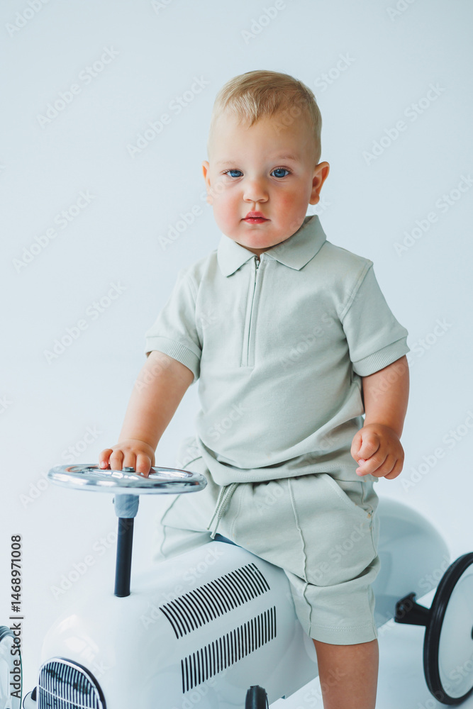 Stylish one-year-old boy in a suit with shorts and a T-shirt rides a car. Toddler boy on a white background with a toy car.