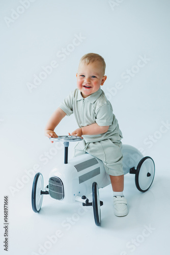 Obraz na płótnie Stylish one-year-old boy in a suit with shorts and a T-shirt rides a car. Toddler boy on a white background with a toy car. w salonie