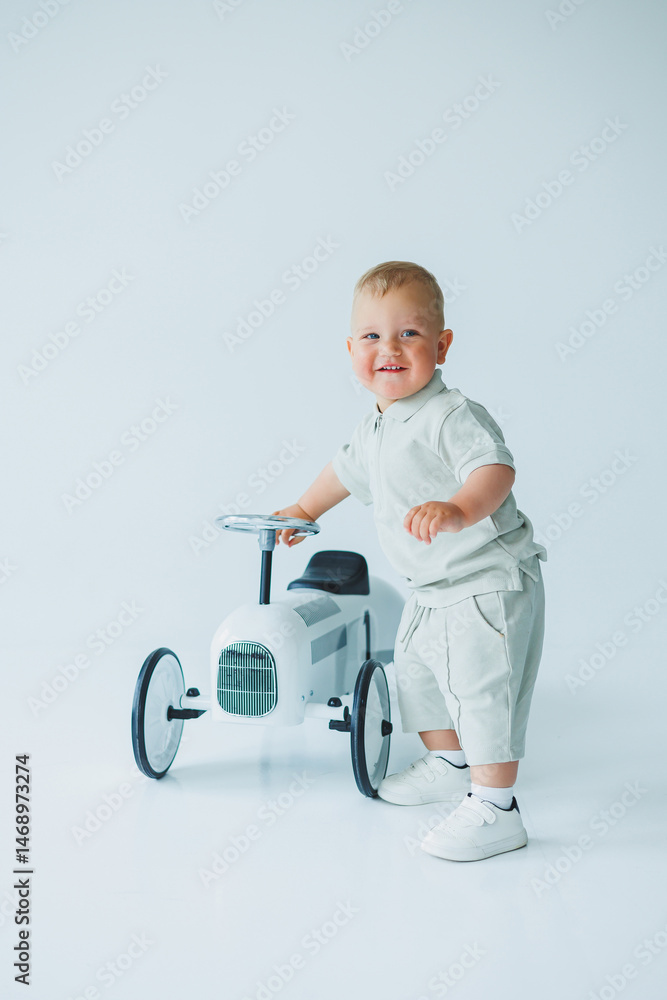 Stylish one-year-old boy in a suit with shorts and a T-shirt rides a car. Toddler boy on a white background with a toy car.