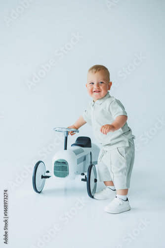 Obraz na płótnie Stylish one-year-old boy in a suit with shorts and a T-shirt rides a car. Toddler boy on a white background with a toy car. w salonie