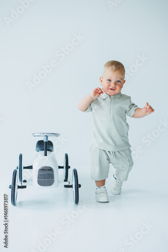 Obraz na płótnie Stylish one-year-old boy in a suit with shorts and a T-shirt rides a car. Toddler boy on a white background with a toy car. w salonie