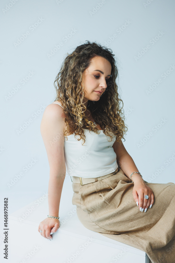 Curly pretty woman sitting in the living room on the sofa and smiling dreamily. Woman in a white top and skirt with a romantic mood sitting in the living room.