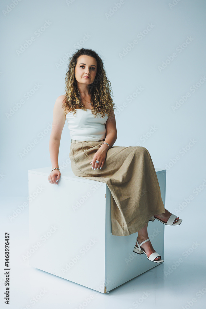 Curly pretty woman sitting in the living room on the sofa and smiling dreamily. Woman in a white top and skirt with a romantic mood sitting in the living room.