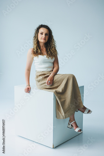 Obraz na płótnie Curly pretty woman sitting in the living room on the sofa and smiling dreamily. Woman in a white top and skirt with a romantic mood sitting in the living room. w salonie