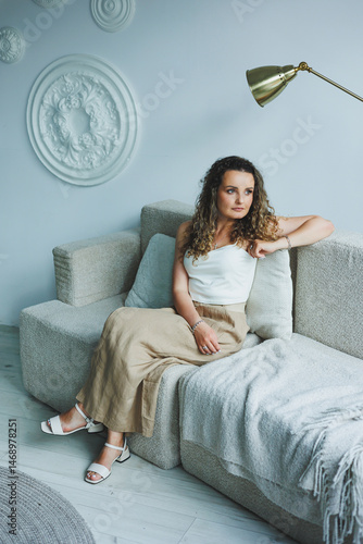 Obraz na płótnie Curly pretty woman sitting in the living room on the sofa and smiling dreamily. Woman in a white top and skirt with a romantic mood sitting in the living room. w salonie