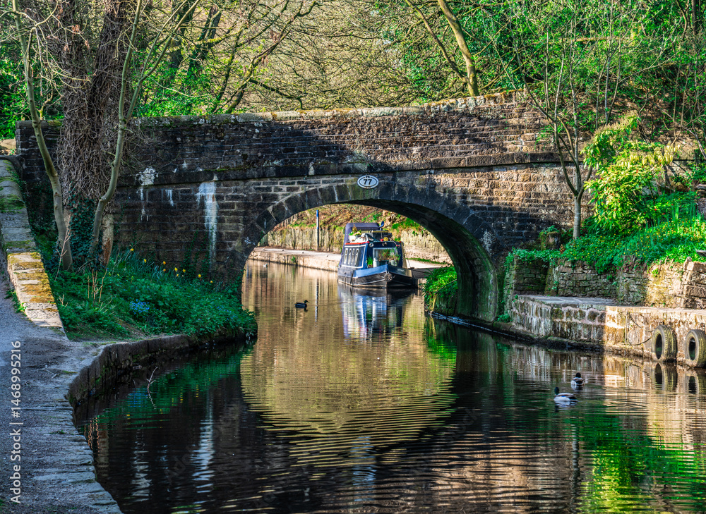 Fototapeta premium Narrow boat moored on canal side with old stone bridge in partial sunlight