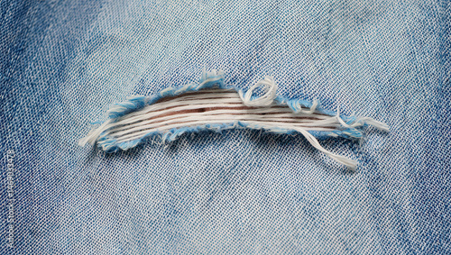 Close-up of torn denim fabric with detailed fringe and visible hole. Light grey blue jean cloth macro texture. Worn, distressed look, ideal for fashion, design projects