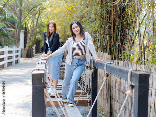 Two beautiful asian women cheerful enjoying sunny day outdoor standing pose on wooden and steel walkway in green park wearing casual outfits smiling look at camera concept friendship weekend lifestyle
