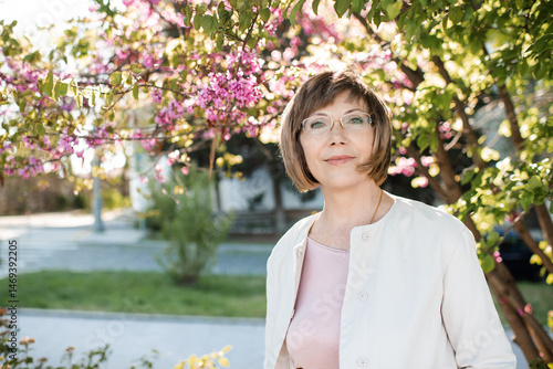 Smiling blonde mature woman 55-60 year old wearing eye glasses posing over flowers in park outdoors close up. Looking at camera. Wellness lifestyle.