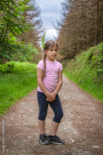 A young girl stands happily on a serene and peaceful forest path, wearing a beautiful floral crown and casual summer attire, surrounded by vibrant lush greenery, Carlingford, Ireland