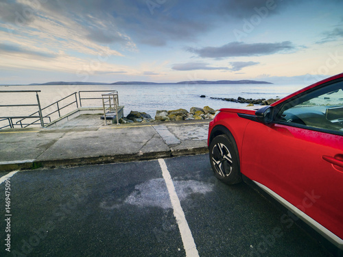 Red car parked in car park with stunning view on nature scenery with ocean and mountains and cloudy sky. Silverstrand area, Galway city, Ireland. Rent motor for a tourist trip concept.
