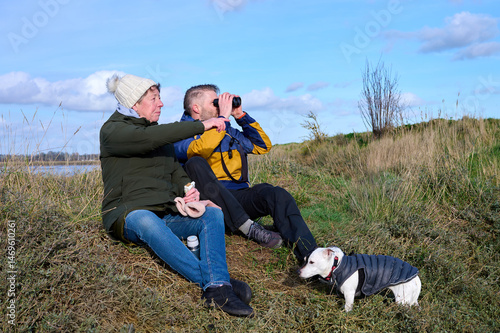 A senior woman and her adult son sit and relax with a binoculars and a snack in a grassy, marsh landscape with their dog on a partly cloudy day.