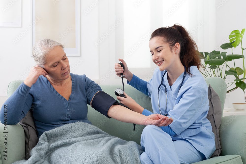 Obraz premium Young friendly smiling nurse or doctor measuring blood pressure of senior elderly woman patient sitting on sofa in medical clinic or at home. Health care concept.