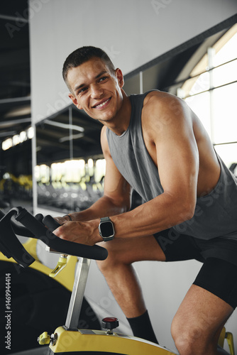 Vertical photo of smiling fit guy at gym exercising on stationary bike. Happy cheerful athlete male training on exercise bike. Young man working out at a class in the modern gym.