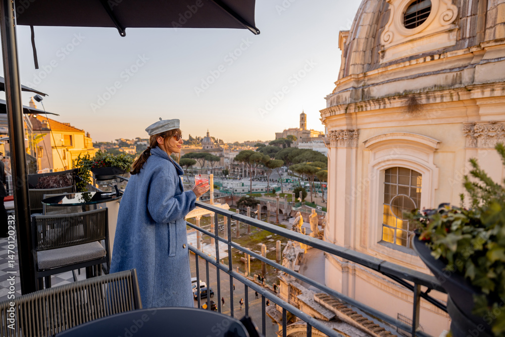 Fototapeta premium Stylish woman with drink enjoys golden hour on a rooftop terrace in Rome. Scenic view over domes, monuments, and ancient ruins