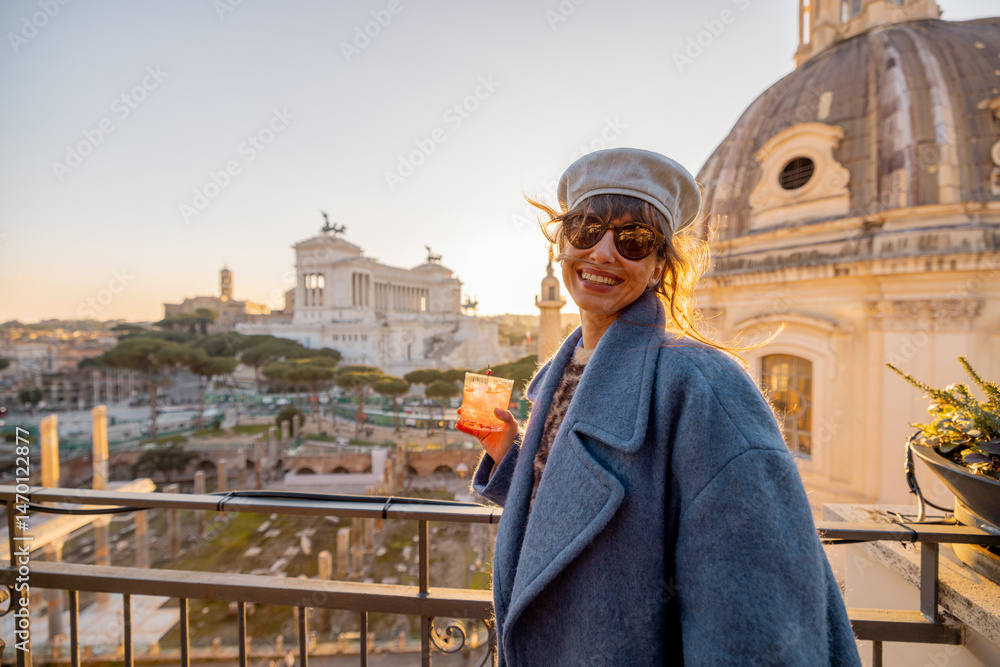 Fototapeta premium Stylish woman with drink enjoys golden hour on a rooftop terrace in Rome. Scenic view over domes, monuments, and ancient ruins