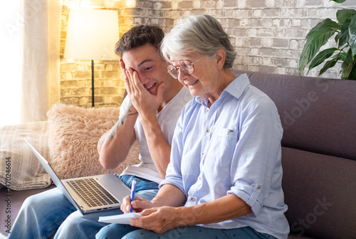 Schilderij op canvas Young adult sitting on couch at home helps his elderly grandmother surf the web on laptop while she takes notes