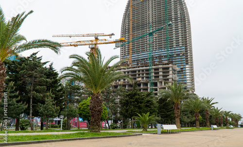 Concept of building a multi-storey city house. View of the building under construction with giant cranes nearby. In front part green park with palm trees and a pedestrian area with benches.