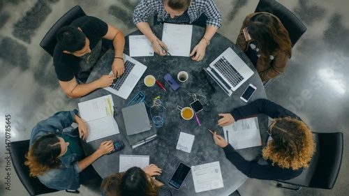 Overhead shot capturing a dynamic team working on laptops, notebooks, and documents at a round table in a modern office. Coffee cups, pens, and smartphones scattered among them.