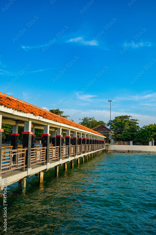 Obraz premium The long bridge supported by the pillar pillar structure and has a fence on both sides and a red roof that stretches over sea water and at the end of the sandy beach area bridge.
