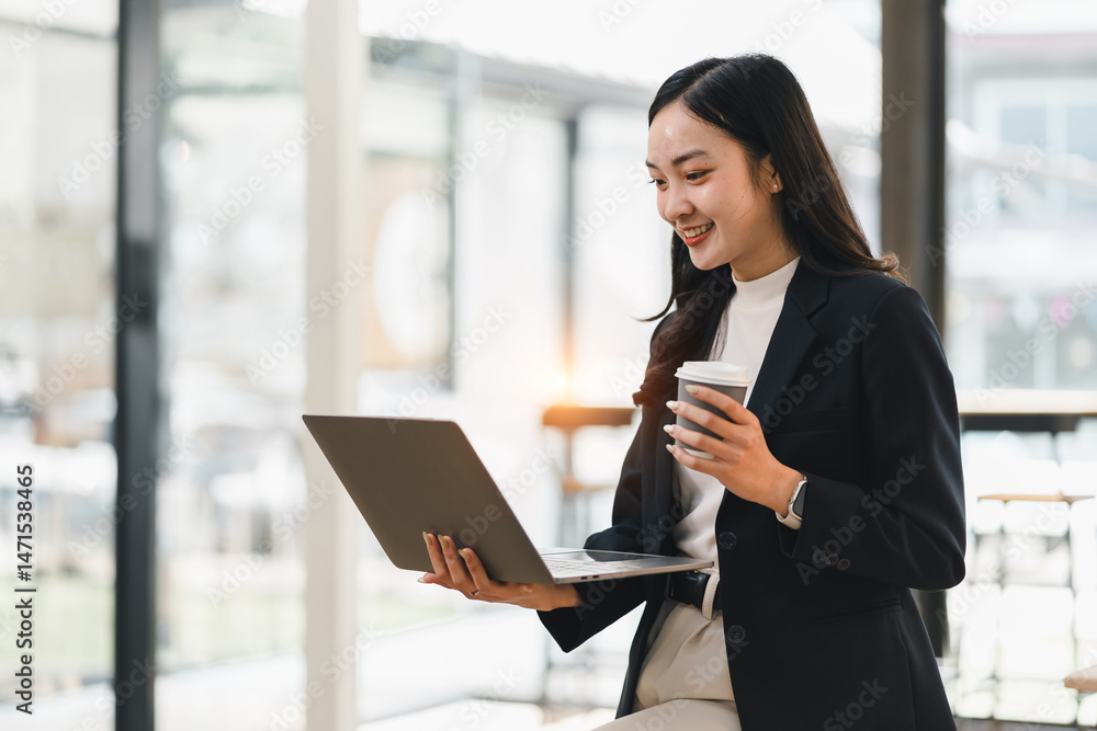 Fototapeta premium Young Asian businesswoman, smiling and holding coffee cup, works on laptop in modern office with bright natural light, expressing confidence and productivity
