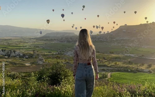 Back view of a solo female traveler walking into a magical sunrise with hot air balloons soaring over Göreme’s valleys. Perfect moment of adventure and discovery.