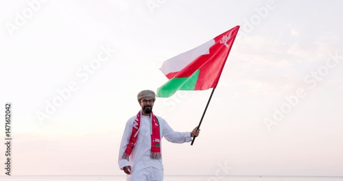 Omani man stands with Omani National Flag, wearing omani traditional dishdasha and Musaar