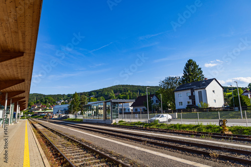 A quiet railway station with tracks extending towards a small village nestled in the hills, featuring modern architecture and a bright, sunny day, highlighting the beauty of countryside travel