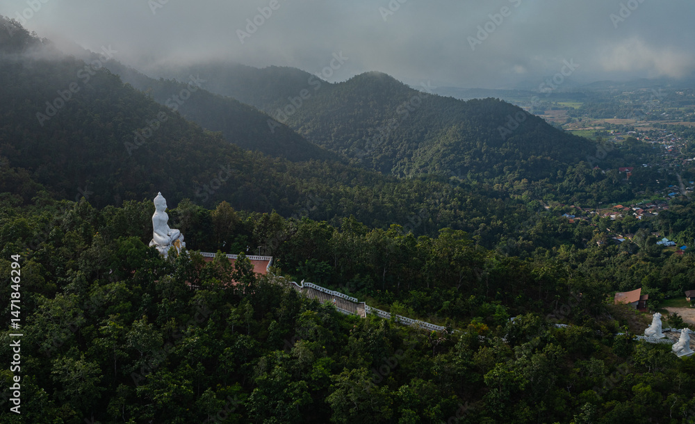 Fototapeta premium A serene aerial view of a large white Buddha statue perched on a forested hill, surrounded by lush greenery and overlooking a peaceful valley with distant villages. The tranquil setting exudes spiritu