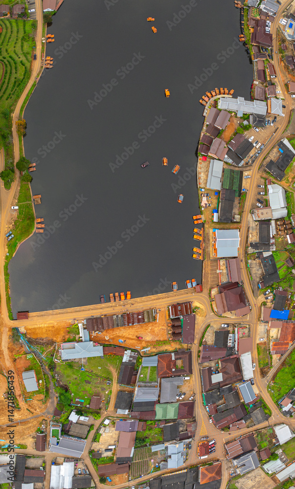 Naklejka premium aerial top view of a lakeside village, showcasing a mix of rustic architecture, lush greenery, and boats scattered across the calm water. A harmonious blend of natural beauty and human settlement..