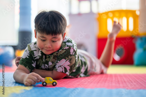 Asian boy lying on colorful playmat playing with toy car focused and calm in indoor playground, learning through play supports child development and imagination, education early childhood activity fun
