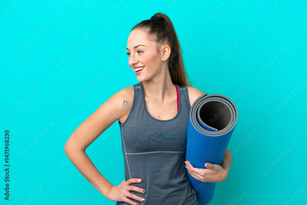 Young sport caucasian woman going to yoga classes while holding a mat isolated on blue background posing with arms at hip and smiling Young sport caucasian woman going to yoga classes while holding a mat isolated on blue background posing with arms at hip and smiling