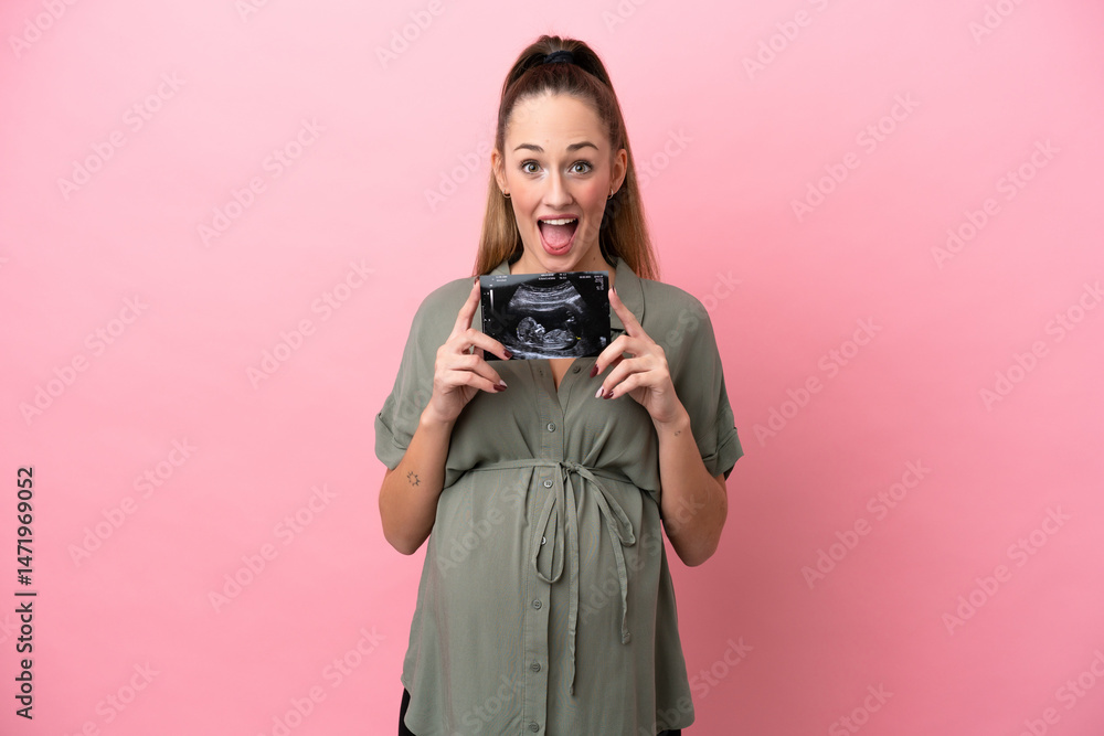 Fototapeta premium Young woman isolated on pink background pregnant and surprised while holding an ultrasound