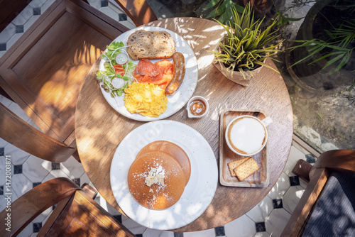 Delicious breakfast with pancakes, sausage, and eggs served on a wooden table surrounded by natural light. A table set in a summer cafe on an open veranda, top view