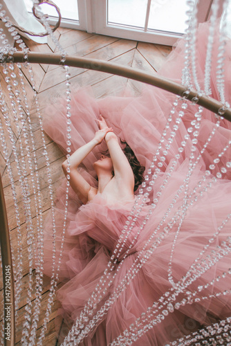 young girl with short black hair in pink tulle dress is lying under crystal chandelier near white window on the wooden floor background at sunny day, camera from top. fashion concept, free space