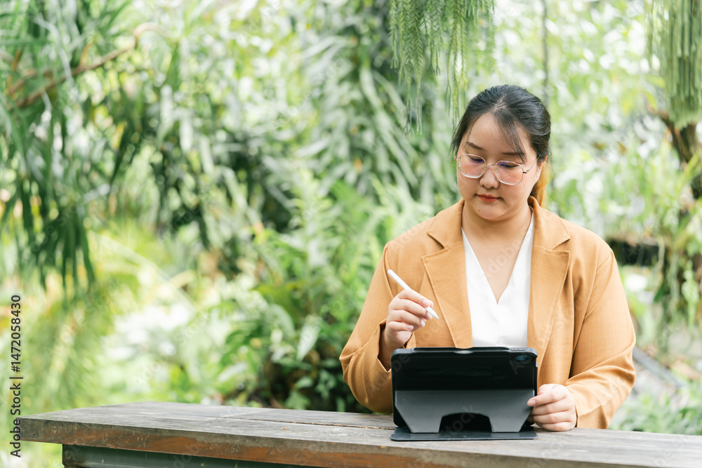 Fototapeta premium Asian Businesswoman A woman writes on a tablet in a lush green outdoor setting, exuding a sense of focus and productivity amidst nature. Female Digital nomad lifestyle