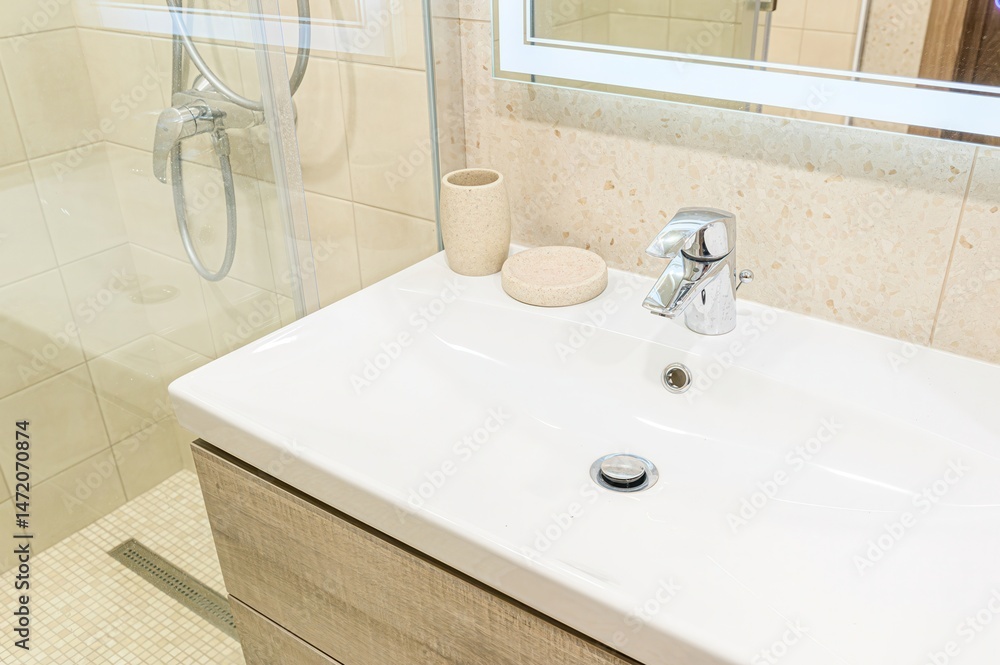 Fototapeta premium clean, well-lit bathroom sink area is shown with a white basin, chrome faucet, and beige accessories. A shower is visible in the background.