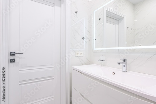 Wallpaper Mural bright, modern bathroom featuring white marble walls, a sleek white vanity, a chrome faucet, and an illuminated mirror. A closed white door is seen on the left. Torontodigital.ca