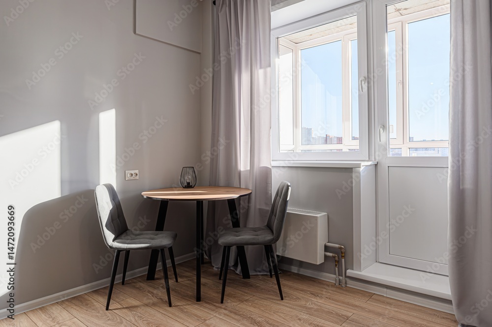 Fototapeta premium Cozy dining area with a round table, two gray chairs, and natural light streaming through a window with light curtains