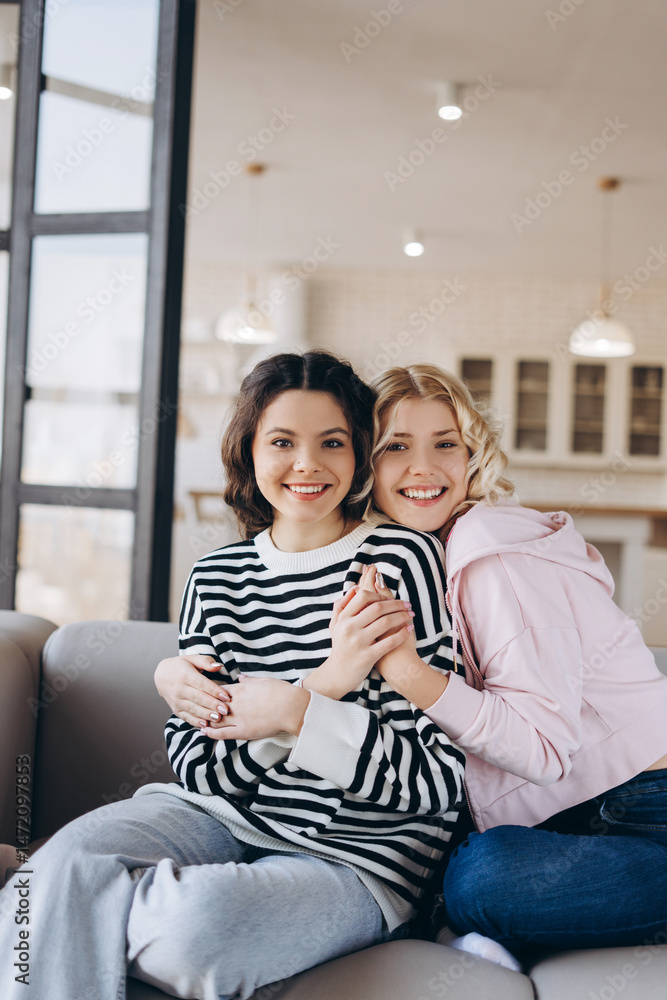 Obraz premium Two amazing girls sitting on the sofa, hugging one another, smiling lovely, looking at the camera