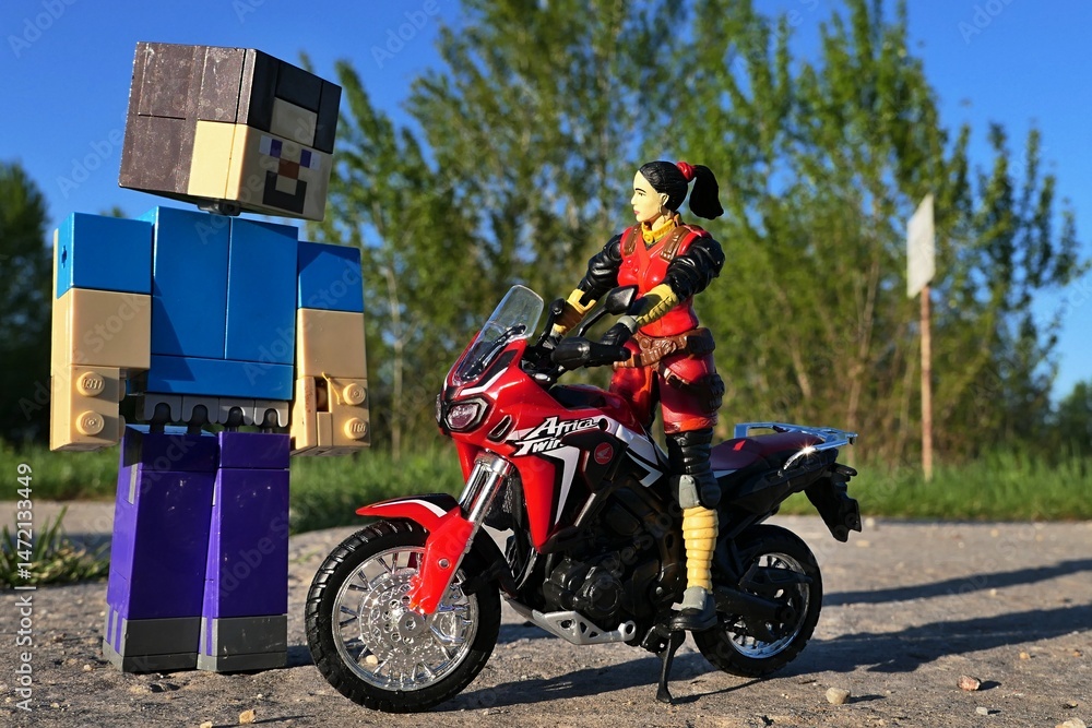 Fototapeta premium GI JOE action figure of female intelligence agent Jinx, version 2004 by Hasbro, standing on red dual sport motorbike Honda Africa Twin, meeting LEGO Minecraft figure of Steve on spring asphalt road. 