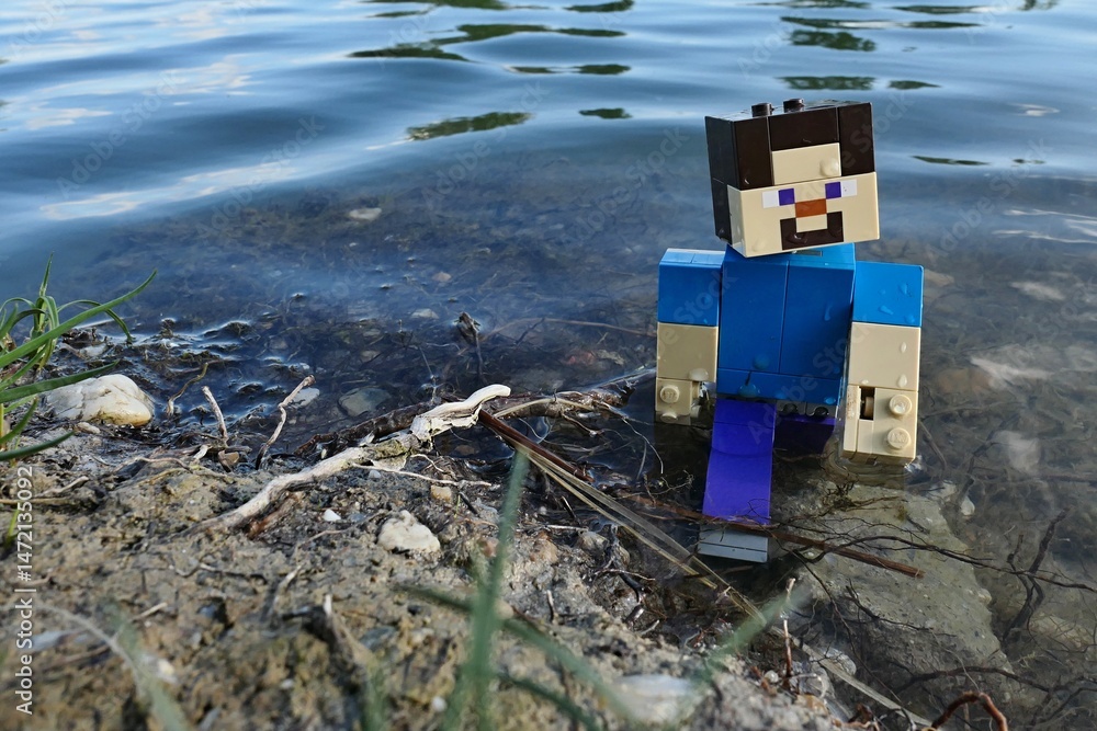 Naklejka premium LEGO Minecraft Steve walking out of shallow water of large spring fish pond, remains of aquatic plants under his feet and reflection of clouds visible.