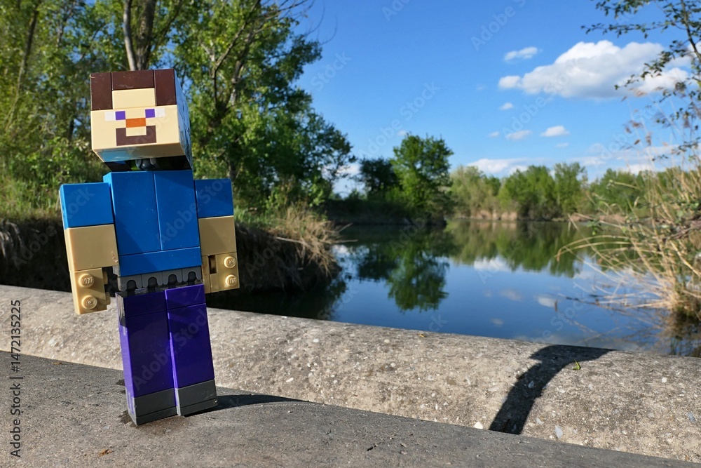 Fototapeta premium LEGO Minecraft action figure of smiling mainc character Steve walking on concrete pipeline bridge across large spring fish pond, broadleaf trees and reed plants visible on bank. Blue skies with clouds