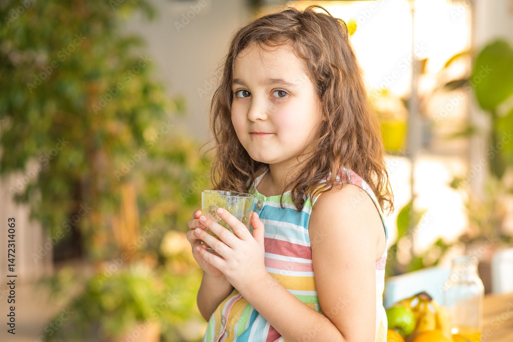 Fototapeta premium Cute little kid girl have breakfast holding a glass of green smoothie at home in the kitchen. Healthy food.