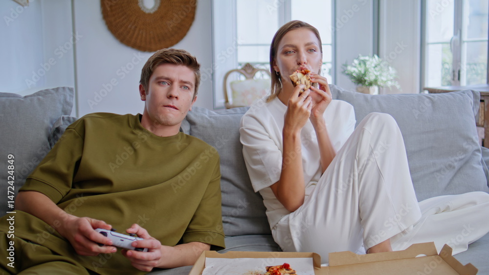 Homie lovers playing console sitting sofa together. Young woman eating pizza