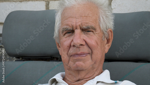 Portrait of elderly gray haired man with deep wrinkles on tanned face looks at camera, sitting on bench near house. Old age in nursing home. Wisdom penetrating look. Loneliness and caring old people