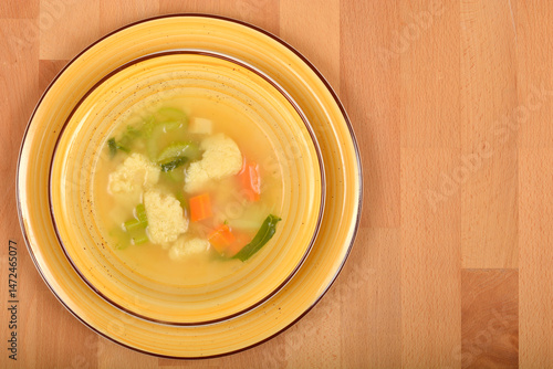 A close-up shows a yellow bowl filled with clear vegetable soup containing semolina dumplings, carrots, and green herbs