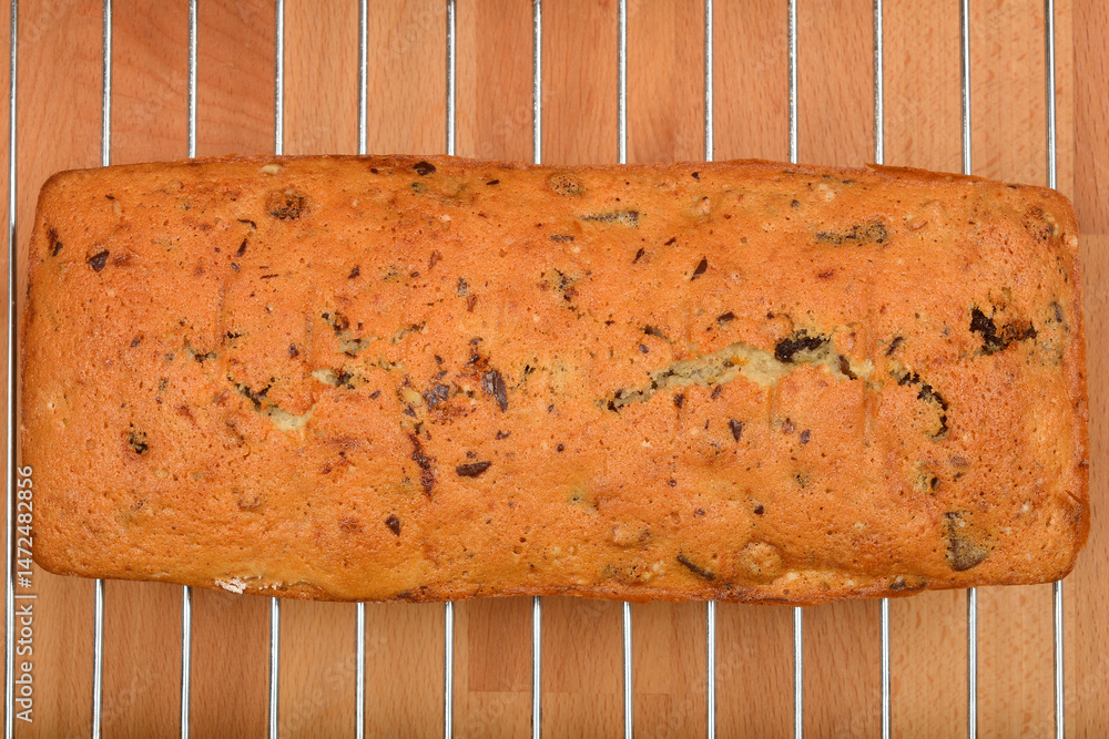 A baked loaf cake with walnuts, raisins, chocolate pieces, and orange zest cooling on a wire rack over a wooden surface