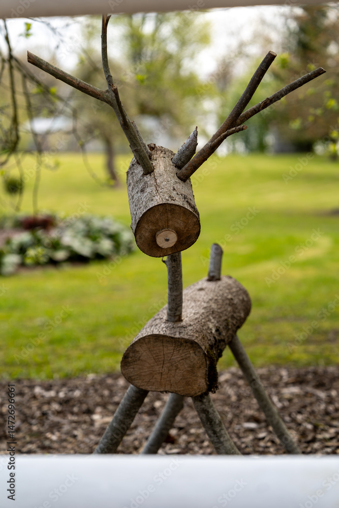 A creative wooden sculpture resembling a reindeer, crafted from tree logs and branches. The sculpture is placed in a natural outdoor setting, with a lush green lawn and colorful flowers in the backgro A creative wooden sculpture resembling a reindeer, crafted from tree logs and branches. The sculpture is placed in a natural outdoor setting, with a lush green lawn and colorful flowers in the backgro