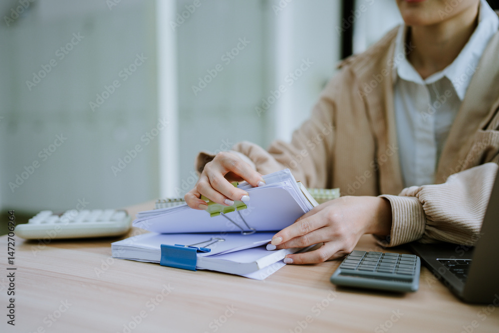Fototapeta premium Hands of businessman hold pen for work in stacks of paper files, information search for business reports and heaps of incomplete documents is achieved on a laptop computer table in a modern office